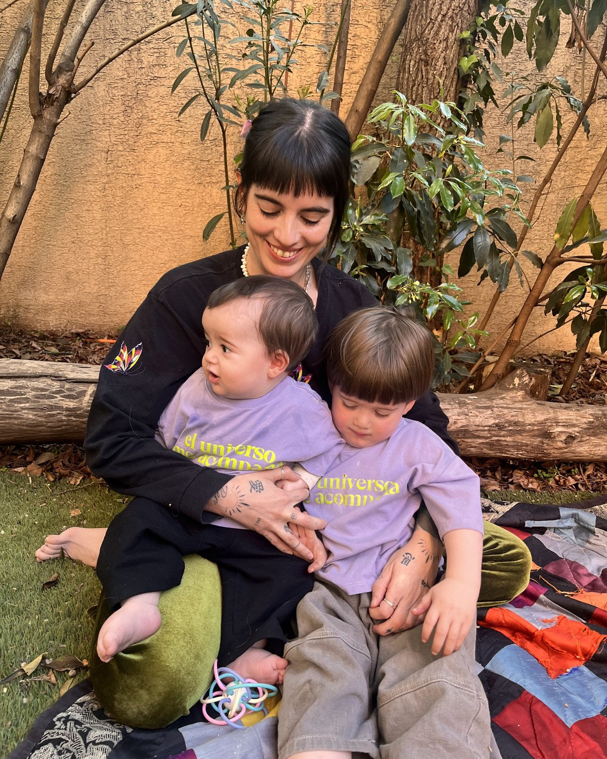 Ali (8 meses) con Polera Universo y Pantal&oacute;n Pantera, ambos talla 1, y Ren (casi 3 a&ntilde;os) con Polera Universo talla 2, sentados en el jard&iacute;n junto a su mam&aacute;