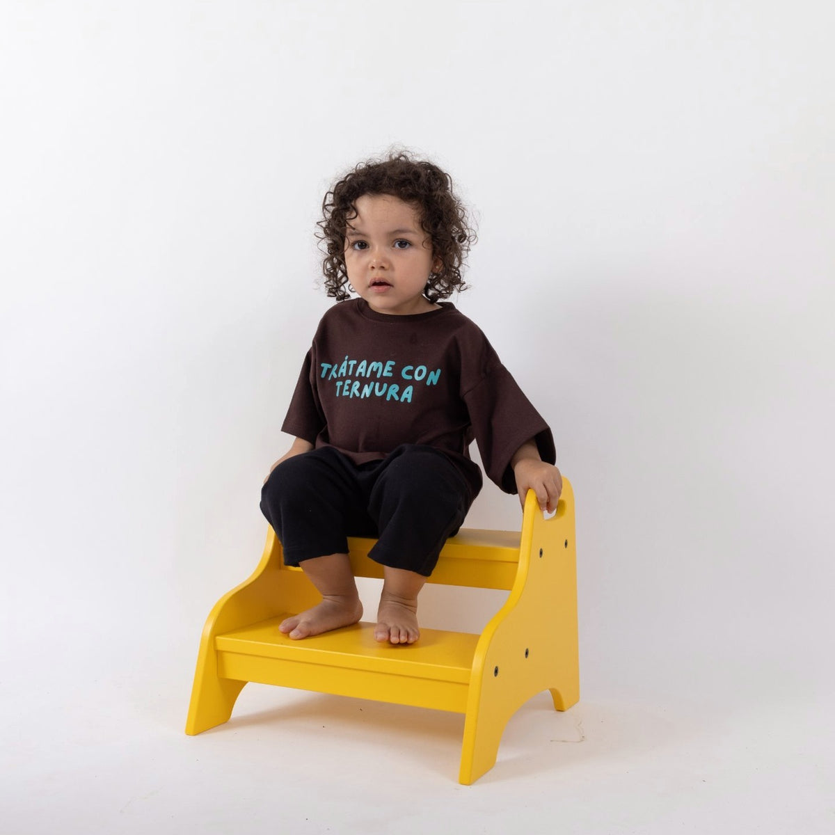 Peque de 2 años sentado en un taburete amarillo con la Polera Ternura café oscuro talla 2 y pantalón negro